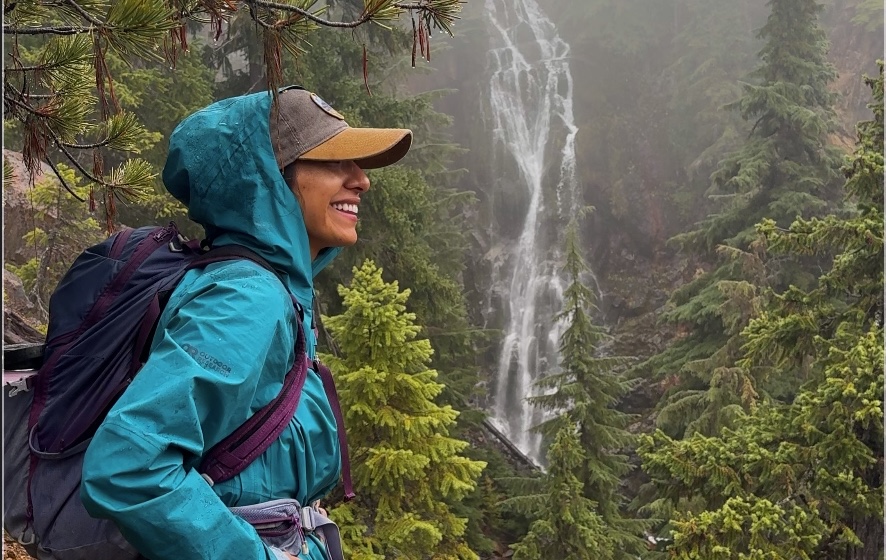 A hiker smiles with Mesatchee Creek Falls cascading in the misty background, surrounded by lush evergreen trees.